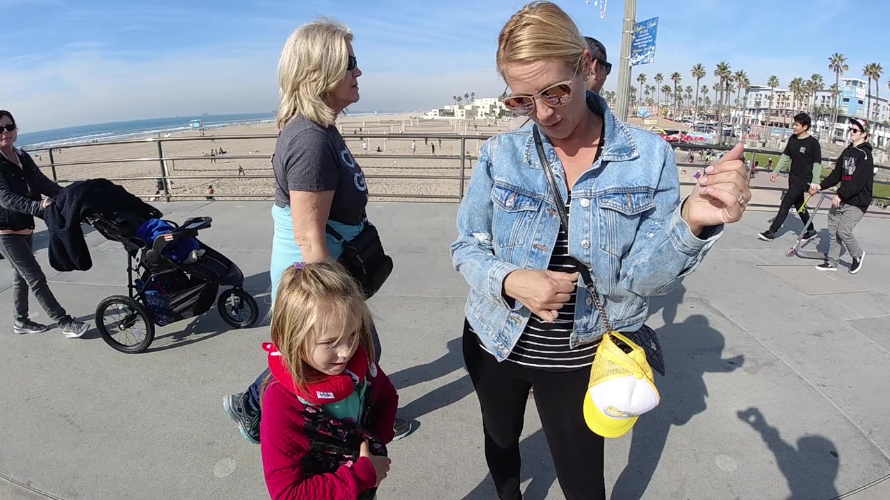 At 0:02 in Huntington Beach, the woman stands near the right foreground with her raised hand visible, the child is near the lower center, and several visitors cross the promenade in front of beach sand and palms.