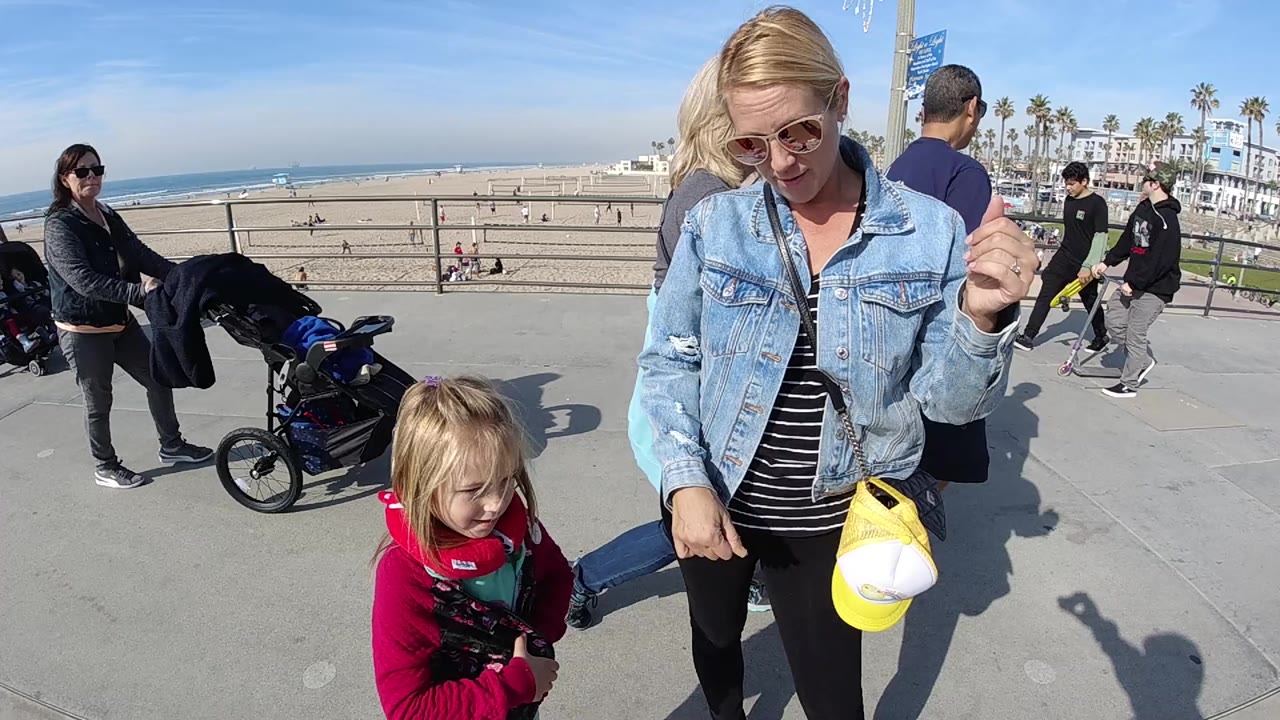 One second in, the child looks down in the foreground as the woman holds a yellow cap, with Huntington Beach sand, ocean, a stroller, and walkers behind them.