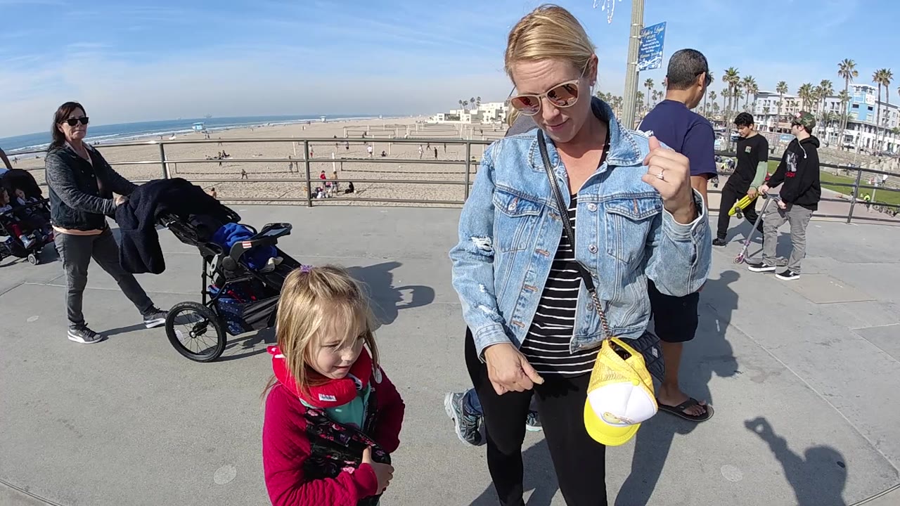 Two seconds in, the woman remains near frame right with the child below her, while several pedestrians pass between the promenade and the Huntington Beach shoreline.