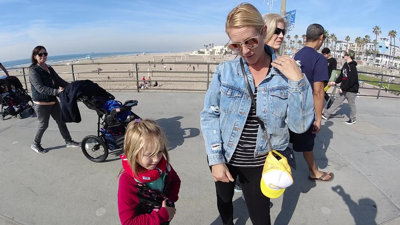 Woman adjusts near her collar while a child stands beside her on a beach promenade.