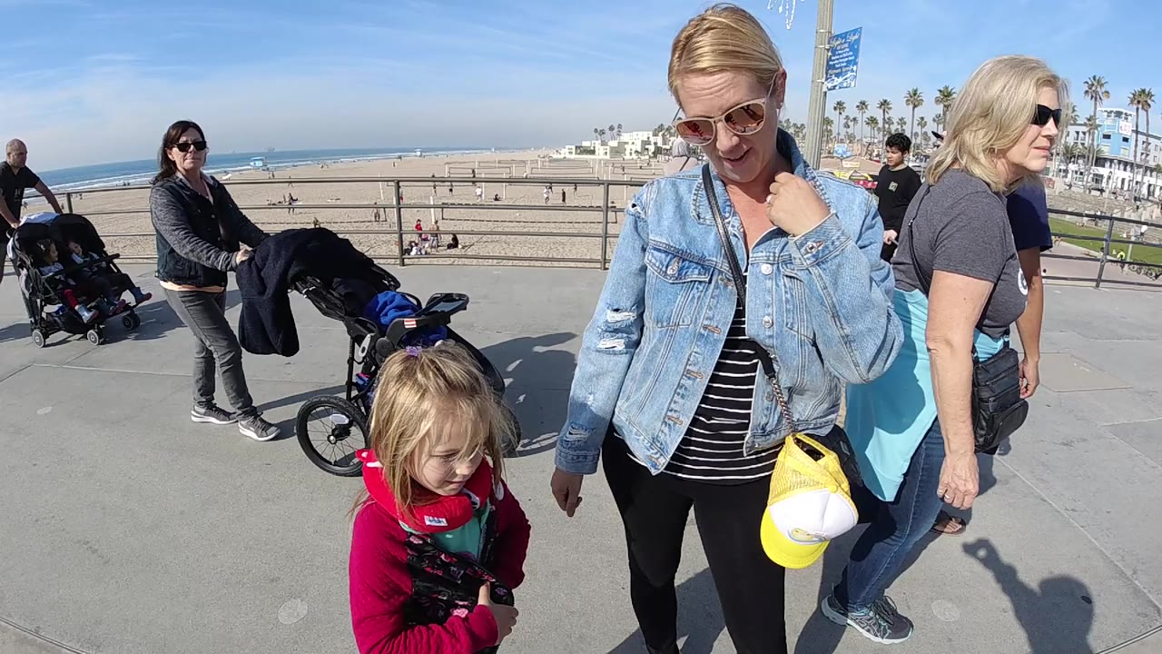 At 0:00 in Huntington Beach, a woman in a denim jacket and sunglasses walks near a small child in red while strollers and beachgoers pass along the oceanfront promenade.