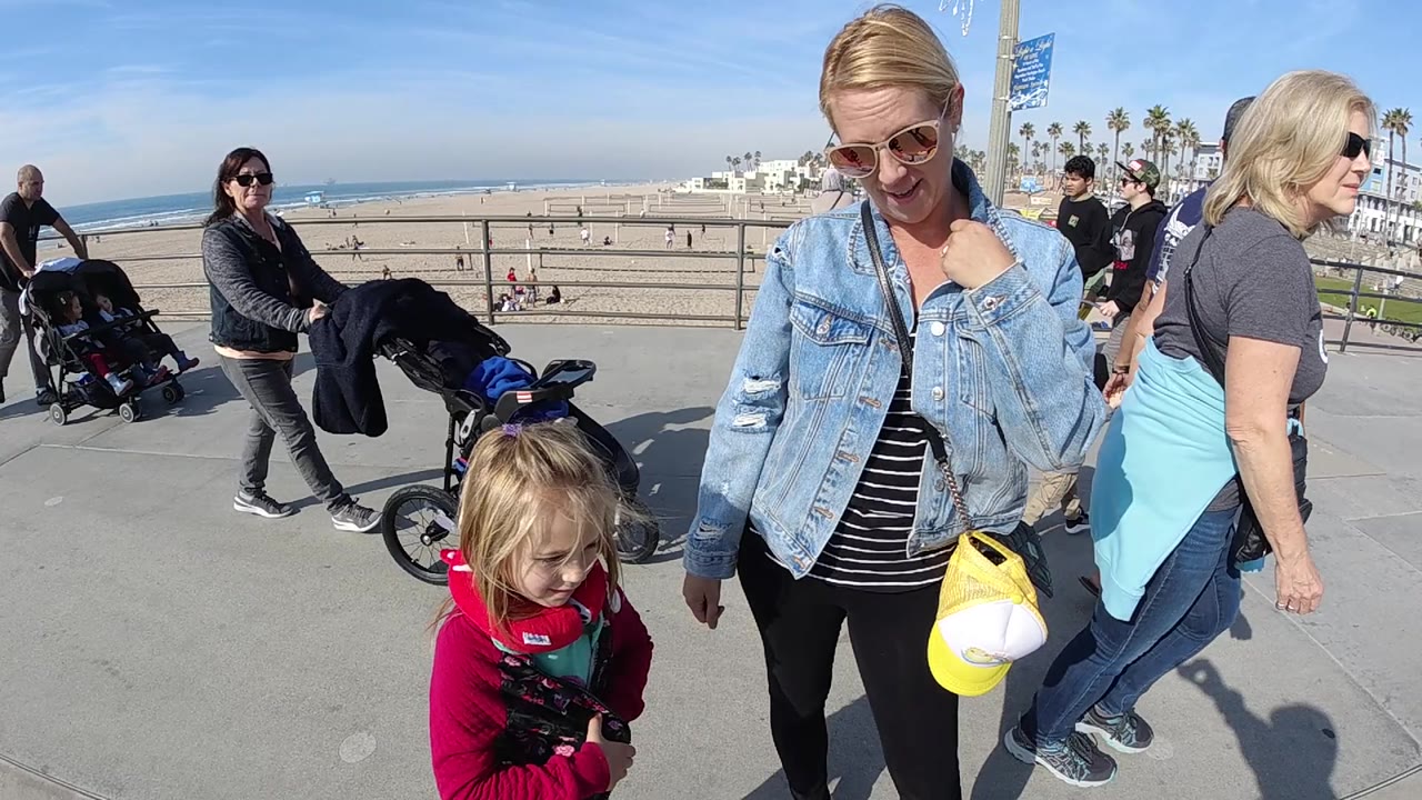 At 0:02 in Huntington Beach, the woman and child stay in the foreground while more pedestrians enter from the right and stroller traffic continues along the promenade.