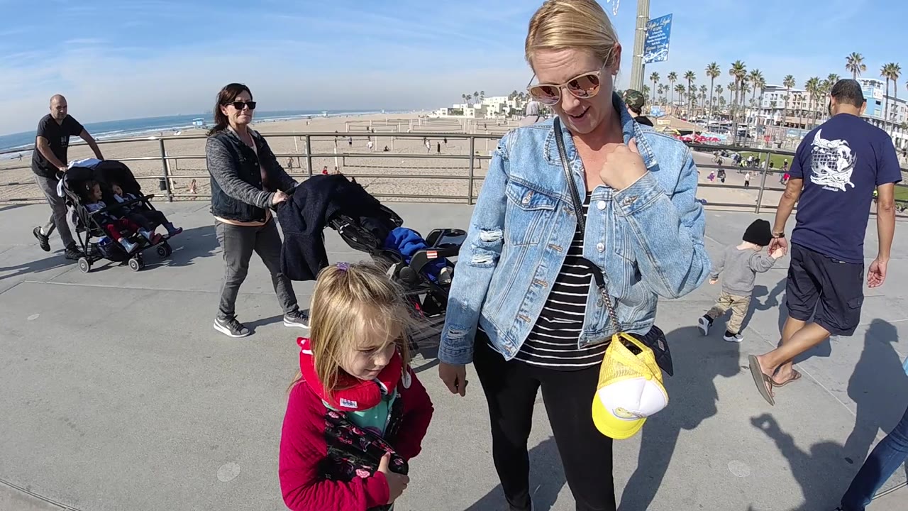 A close foreground view shows the woman holding a yellow cap beside the young girl, with a man and child walking past and Huntington Beach sand and surf in the background.