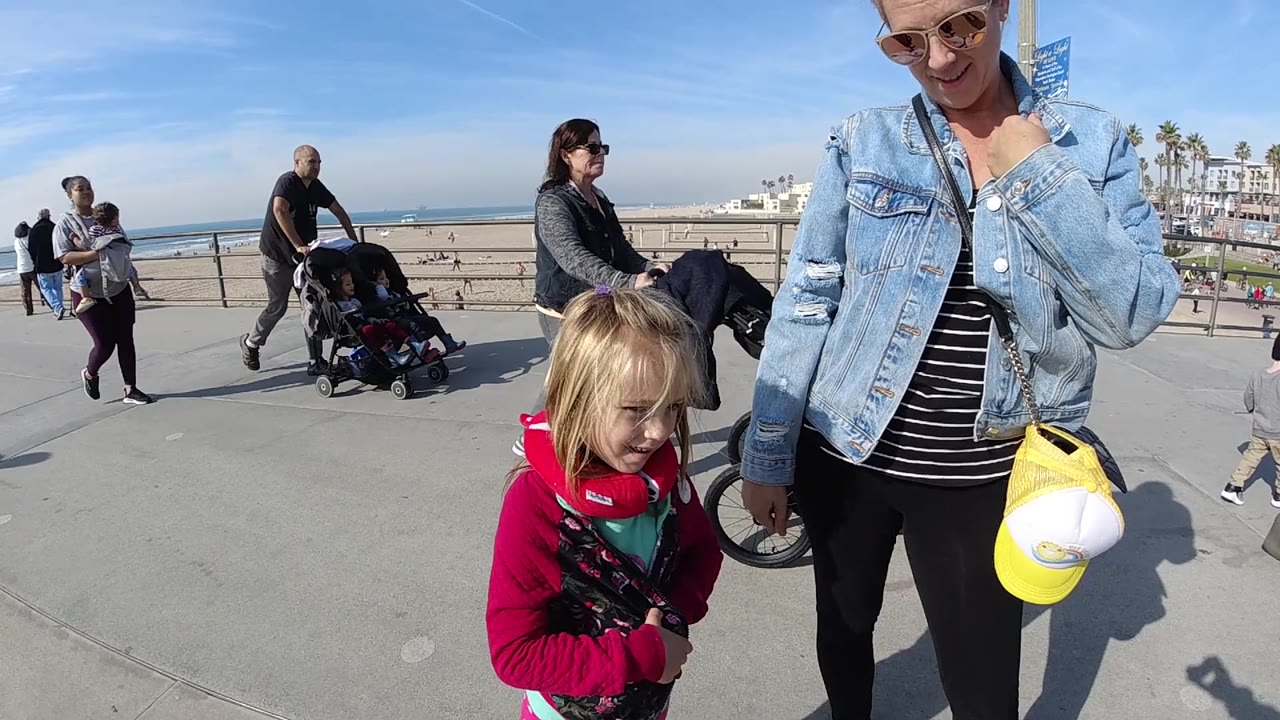 In Huntington Beach, the woman in a denim jacket holds a yellow and white cap at her side as the child stands close by, with beachgoers, strollers, sand, and ocean visible in bright daylight.