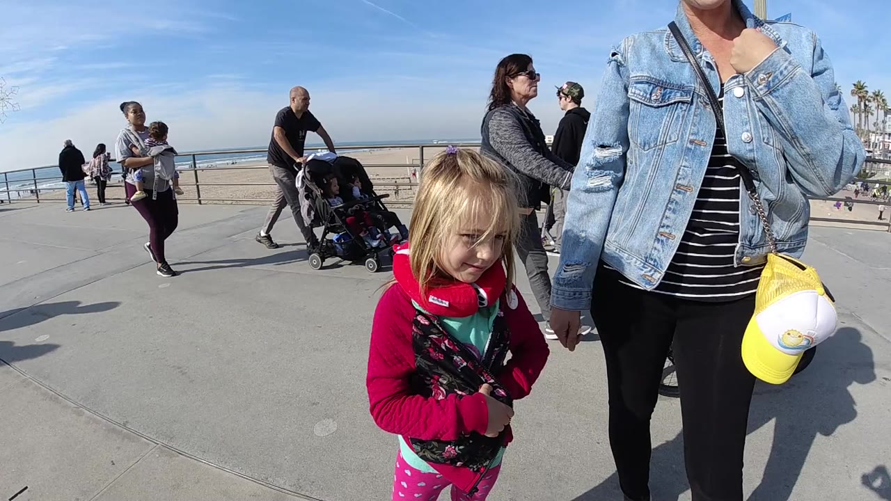 The young girl in pink remains in the foreground beside the adult, while a man with a stroller and other walkers pass along the Huntington Beach oceanfront path.