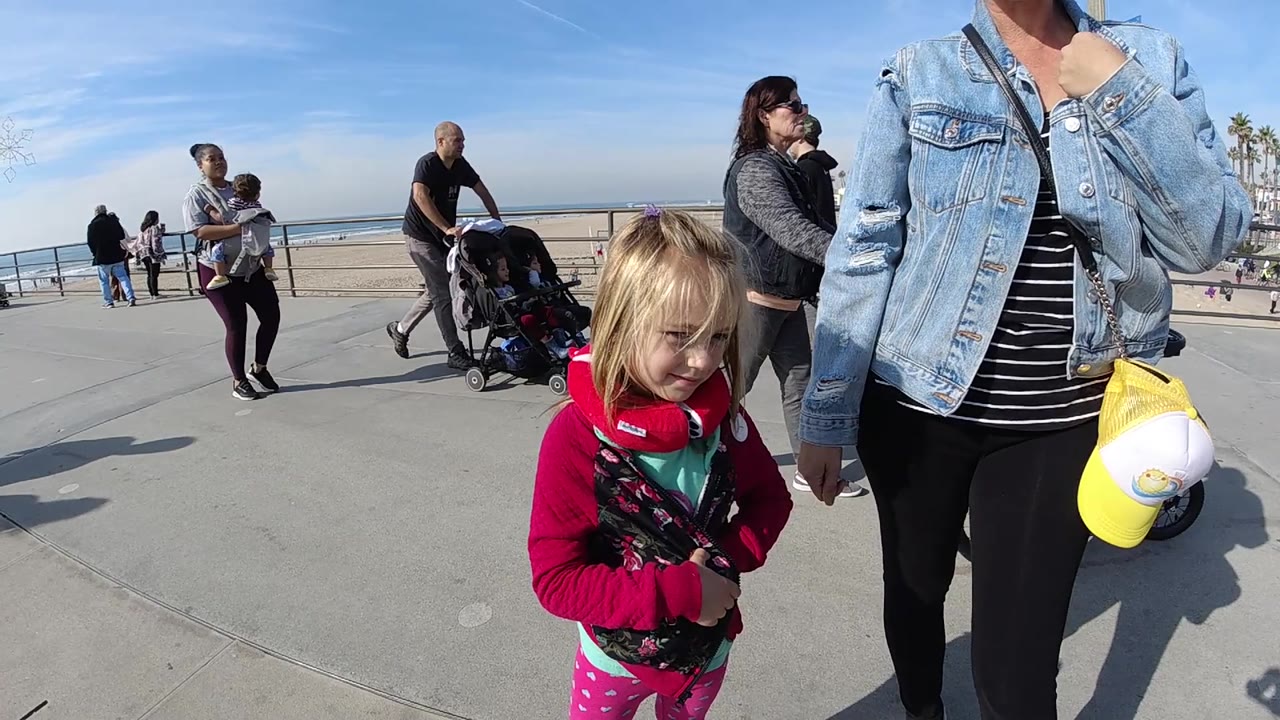 A close foreground view shows the child looking downward beside the adult at Huntington Beach, with several people, a stroller, beach railing, and ocean horizon in the background.