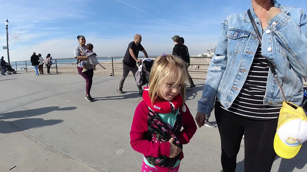A blonde child in a red hoodie stands near an adult in a denim jacket on a Huntington Beach promenade, with pedestrians, railing, sand, ocean, and blue sky behind them.