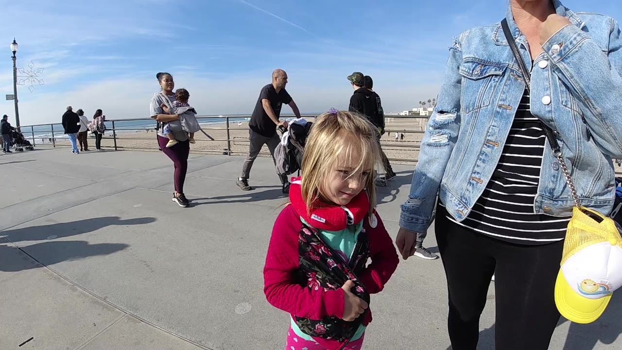The child remains low in the foreground on the Huntington Beach walkway while a woman carrying a child and other pedestrians move near the ocean railing in the background.