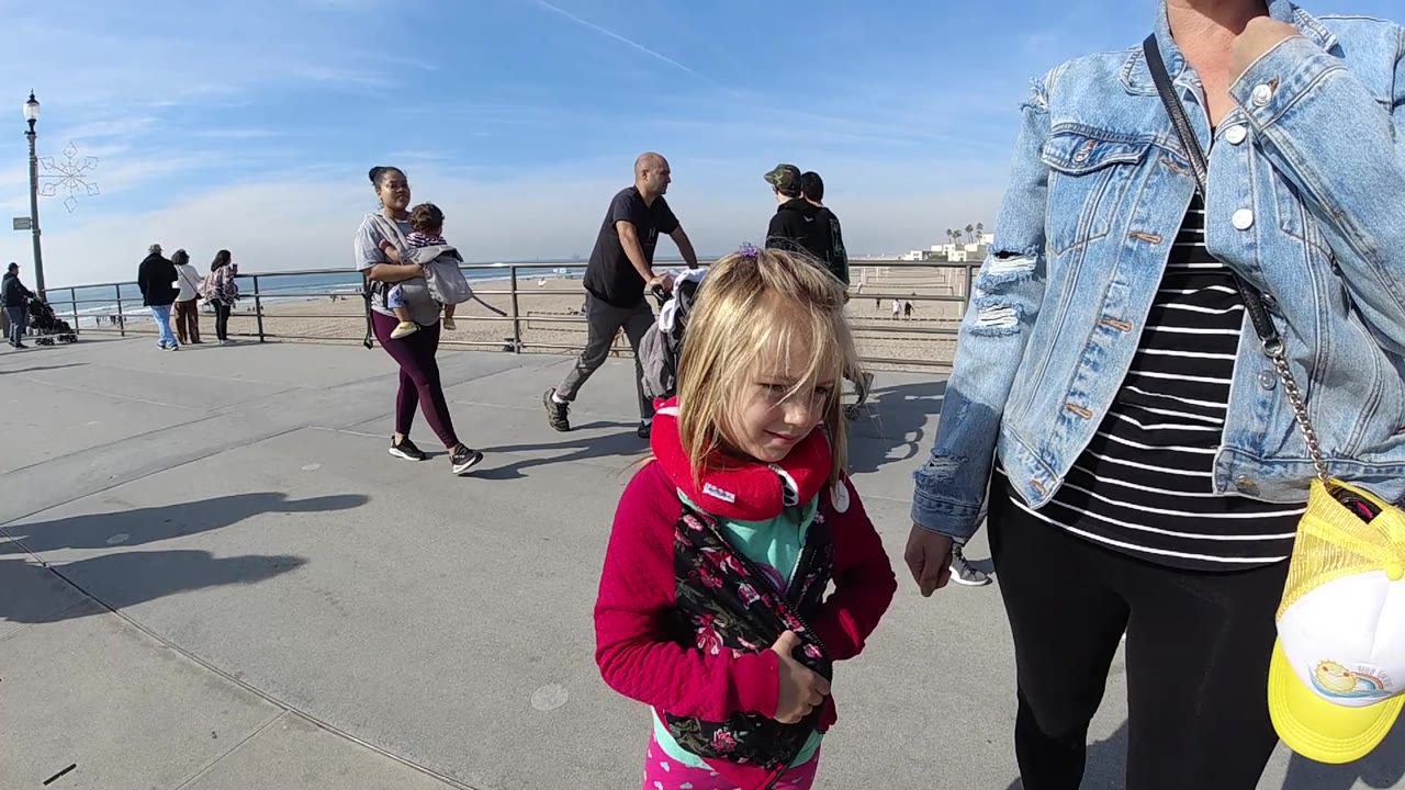 A cropped adult in a denim jacket stands beside the blonde child on the Huntington Beach promenade, with a bald pedestrian, stroller, railing, beach, and ocean visible behind them.