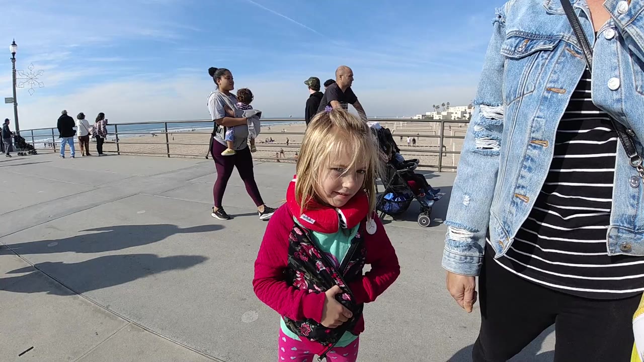 Girl in red outerwear faces the camera beside a cropped adult on a bright coastal walkway.