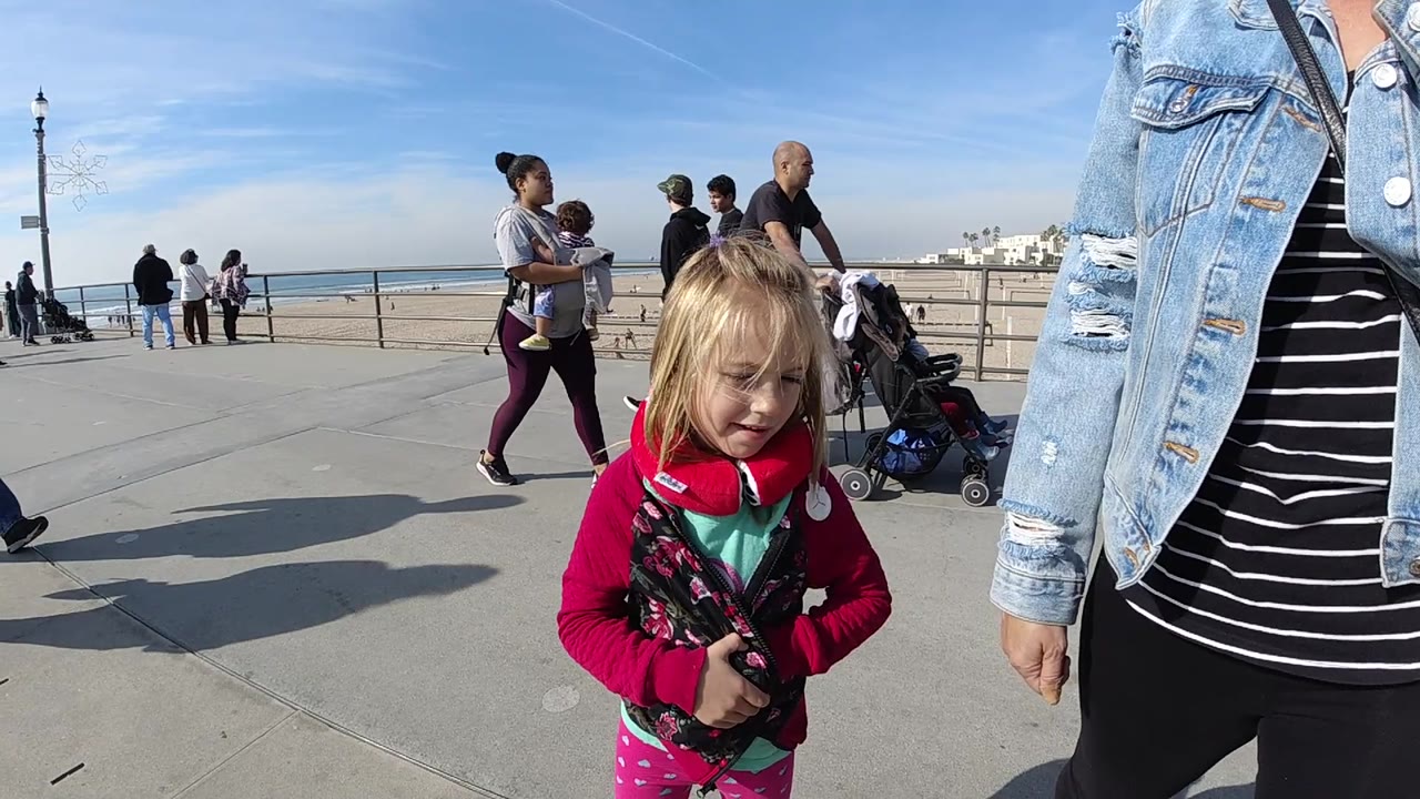 At 0:02 in Huntington Beach, California, the young girl stands slightly right of center with her hands together while several pedestrians and strollers move through the midground near the railing.