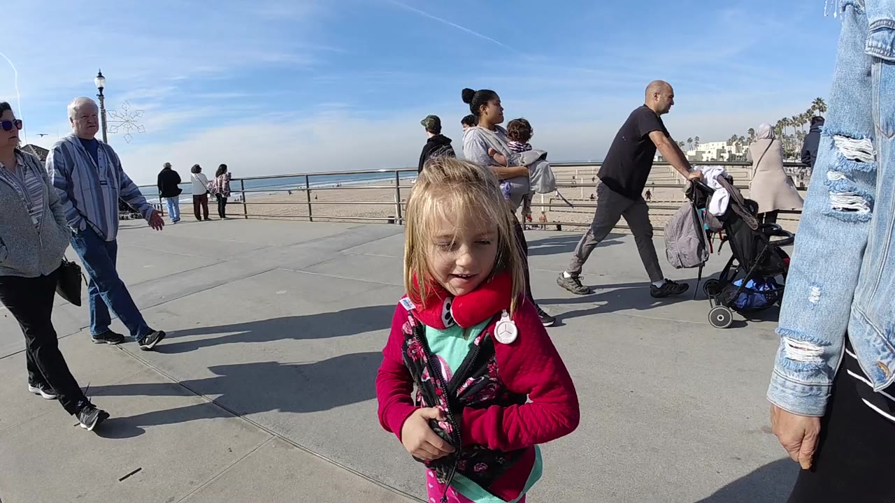 Young blonde girl in a red jacket and red headphones stands near the foreground on a Huntington Beach sidewalk while adults, a stroller, beach sand, ocean, and blue sky appear behind her.
