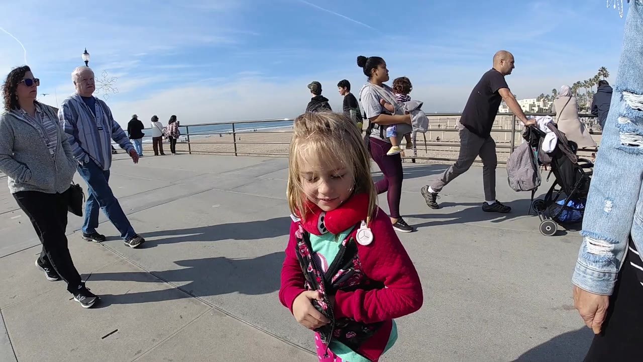 The girl looks downward in the foreground as an older couple, a woman carrying a child, a man with a stroller, beach railing, sand, and ocean remain visible in bright Huntington Beach daylight.