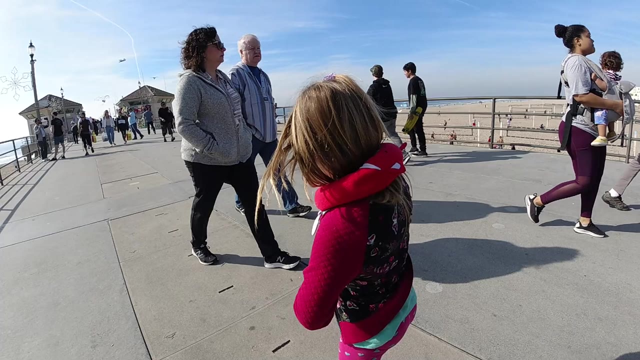 At Huntington Beach, California, a young girl in a red hooded top stands foreground right on the pier boardwalk while adults pass nearby and the beach and ocean sit beyond the railing.