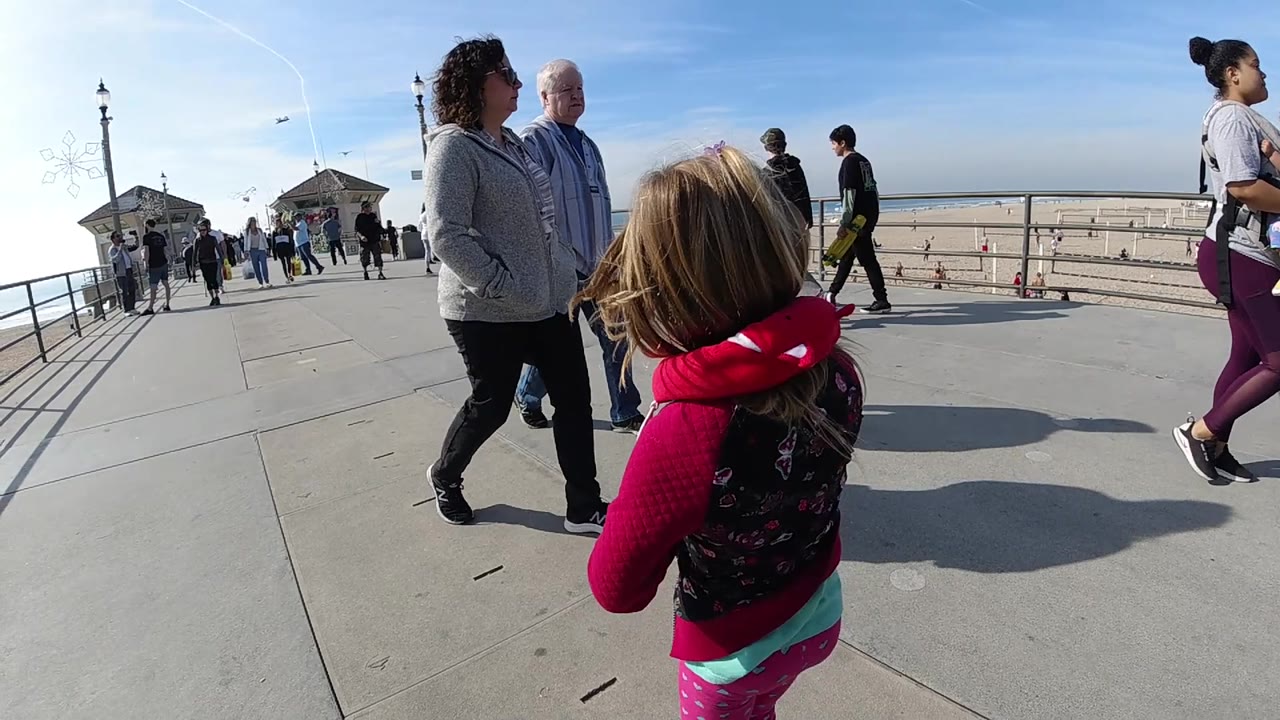 At Huntington Beach, California, the girl remains near the right side of the frame as two adults stand close by, with a pier railing, sandy beach, and ocean horizon extending across the background.