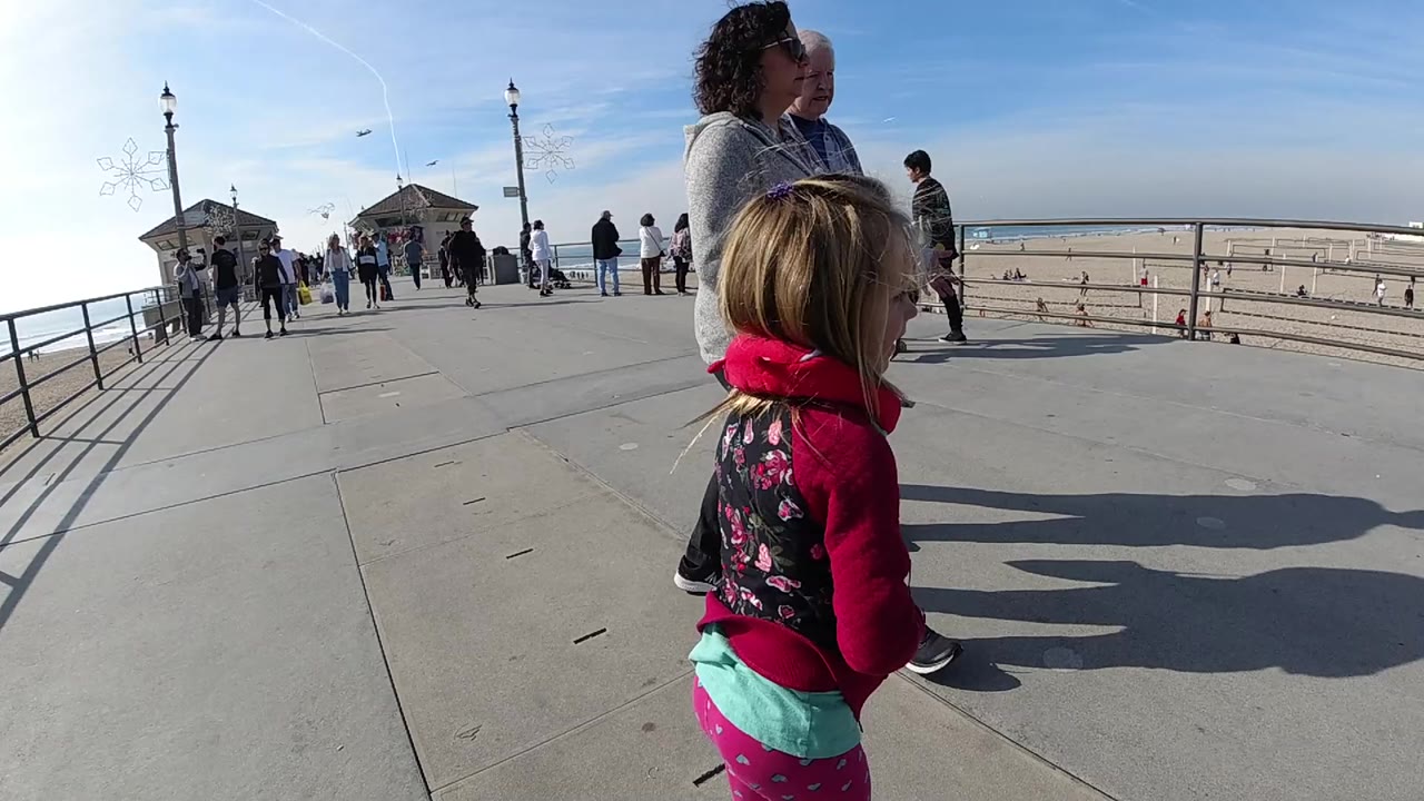 A young girl in a red hoodie walks near an adult woman on a Huntington Beach boardwalk with railings, sand, beach visitors, pavilion structures, and blue sky behind them.
