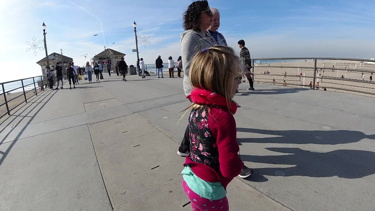 The girl and woman move farther across the Huntington Beach boardwalk as pedestrians gather near the railing and small beach pavilion in the background.
