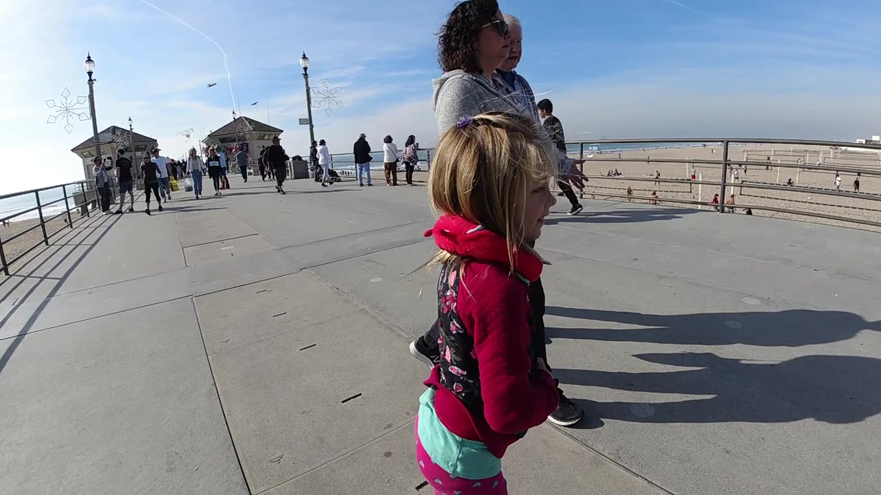 A child in a red hoodie is near the right foreground while the adult woman walks beside her on the Huntington Beach boardwalk, with sand and railings stretching behind them.