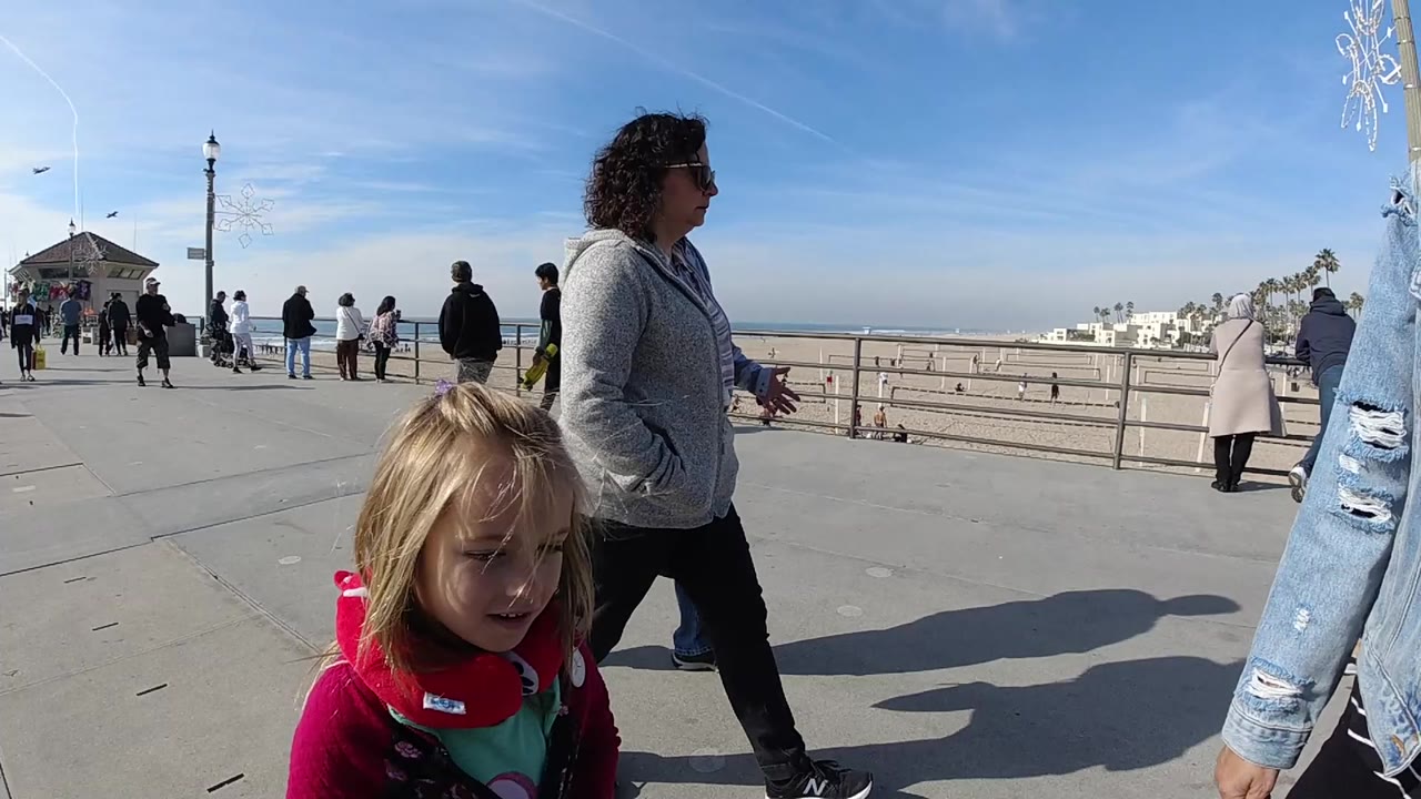At Huntington Beach, a blonde child stands low in the foreground while a woman in a gray hoodie and other pedestrians occupy the boardwalk beside the beach railing.