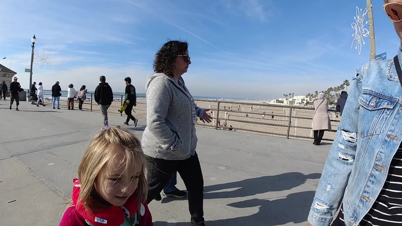 At Huntington Beach, a woman in a gray hoodie stands in profile while a child is partly visible below her and several people move along the railing facing the sand and ocean.