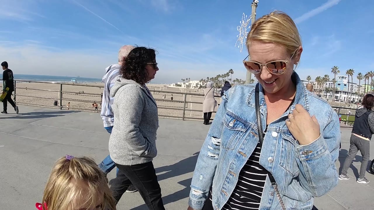 Woman looks down in sunglasses beside a beach railing as people walk behind her.