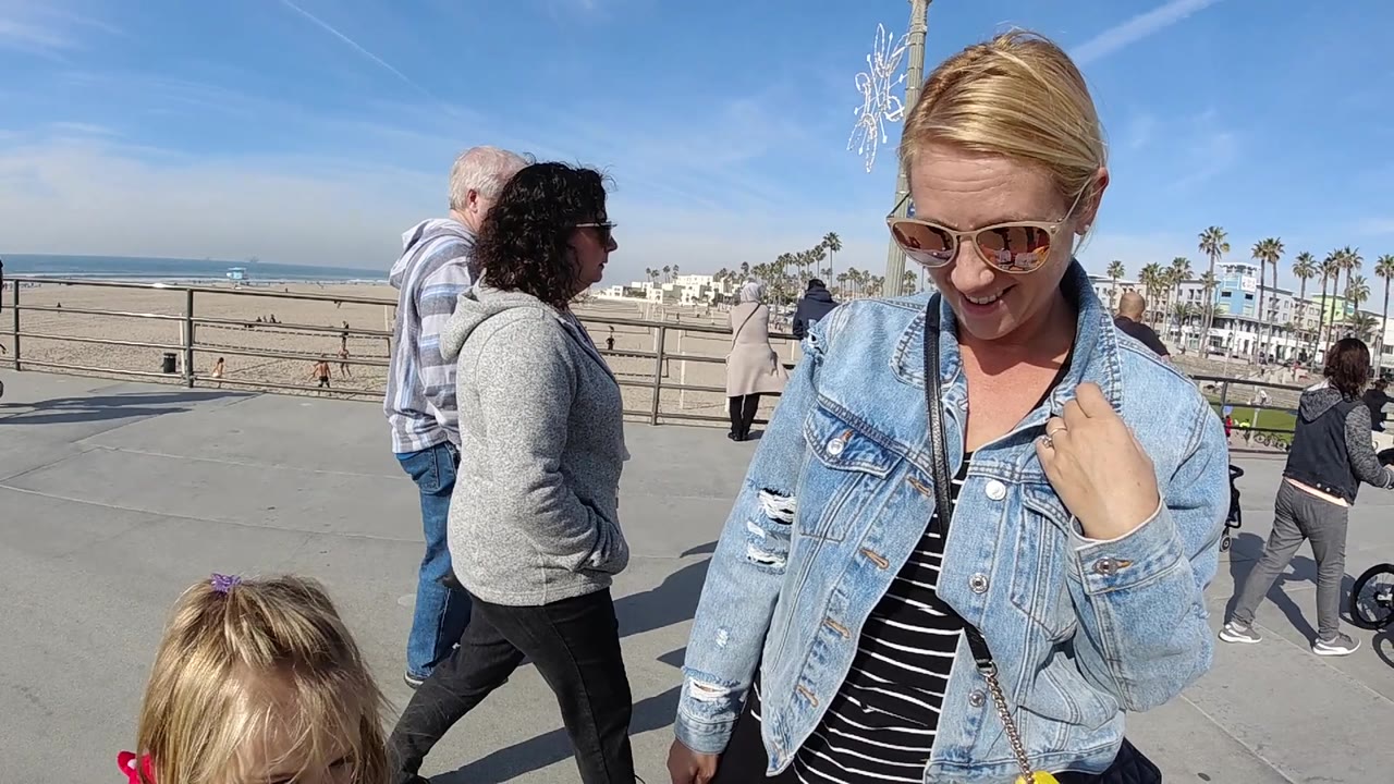 At 0:02 in Huntington Beach, the woman remains close on the right edge while the beach railing, open sand, ocean horizon, palm trees, and multiple pedestrians are visible in hard midday light.