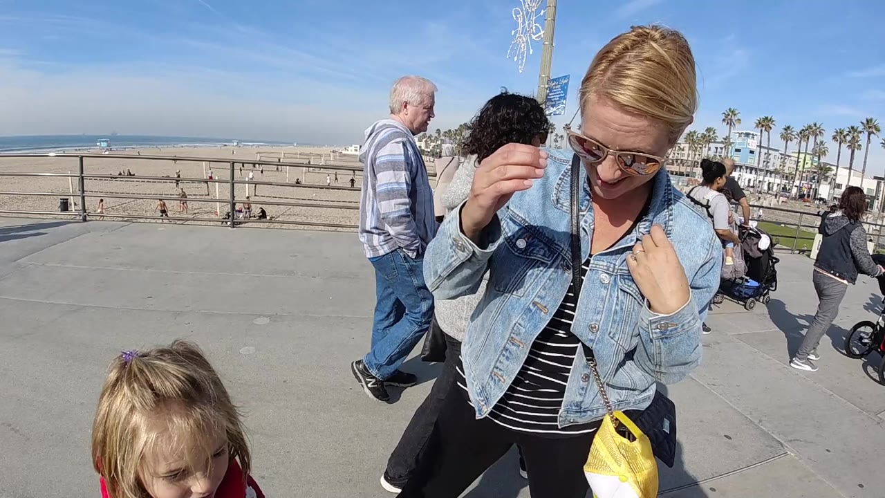 At Huntington Beach, California, the woman touches her sunglasses while the young girl is partly cropped at the bottom and the pier sits beyond the sand.