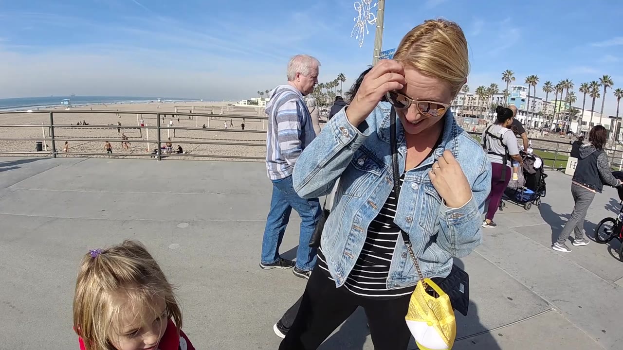 At Huntington Beach, California, the woman raises one hand to her hair beside the cropped child, with a man and other walkers crossing the promenade behind them.
