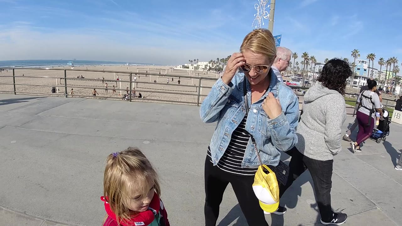 In Huntington Beach, a woman in a denim jacket and sunglasses stands near a small child on a concrete overlook, with beach sand, railings, ocean, and palm trees in the background.