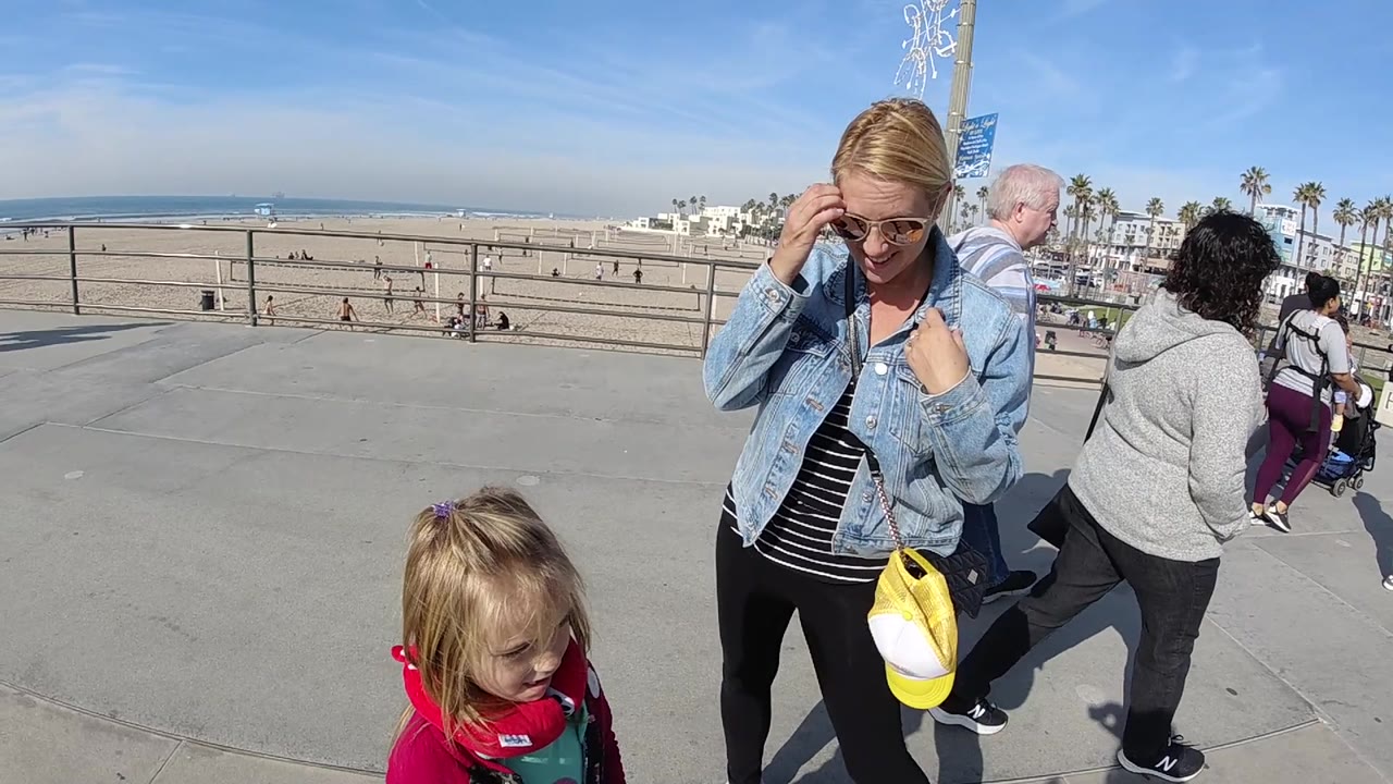The woman stands to the right of the frame as the child looks downward near the lower edge, with beachgoers, railings, sand, ocean, and palm trees visible in Huntington Beach.