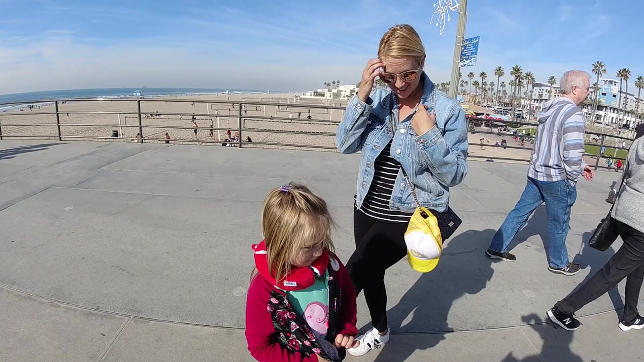 At 0:02 in Huntington Beach, the woman looks down while holding her sunglasses, the child stands lower in frame, and an older pedestrian passes behind them near the beach railing.