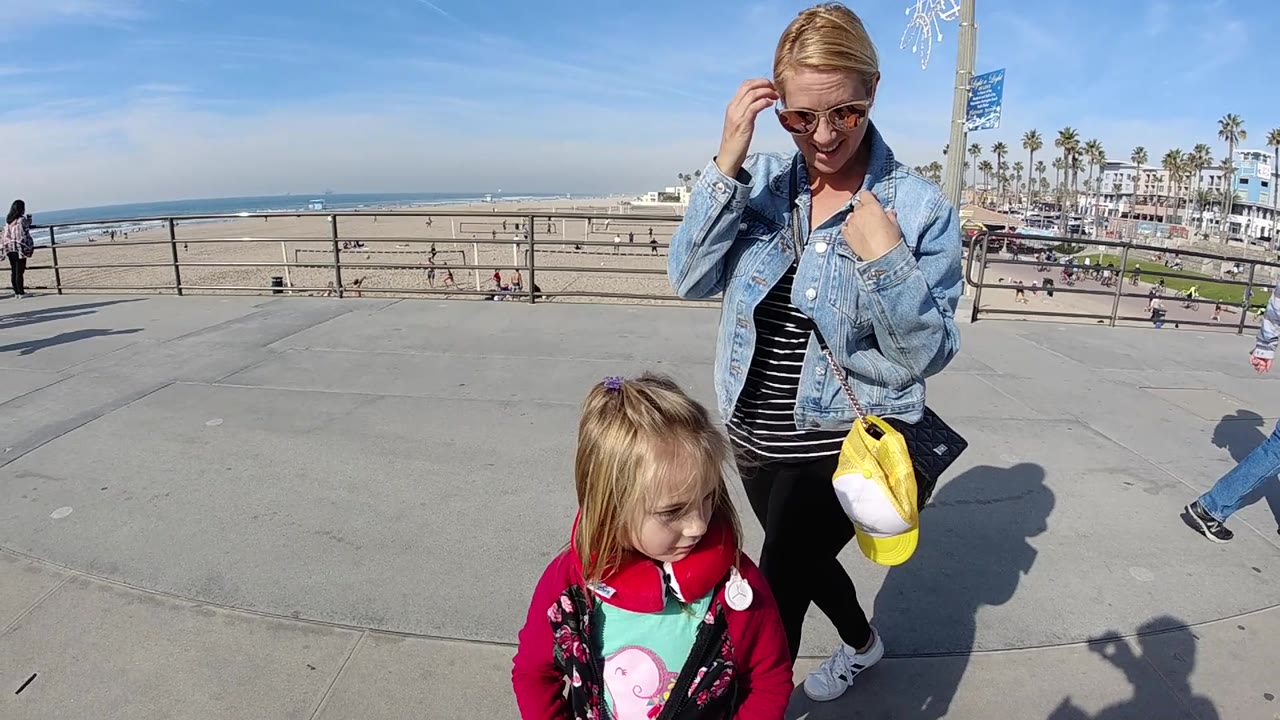 At Huntington Beach, the woman looks downward with one hand near her collar while the child passes lower in frame and the ocean, sand, railings, and palm-lined street remain visible.