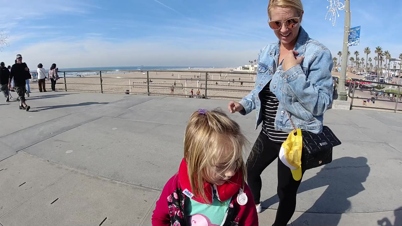 In Huntington Beach, a blonde woman in sunglasses stands near a young blonde child on a concrete promenade, with beach railing, sand, ocean, and pedestrians in the background.