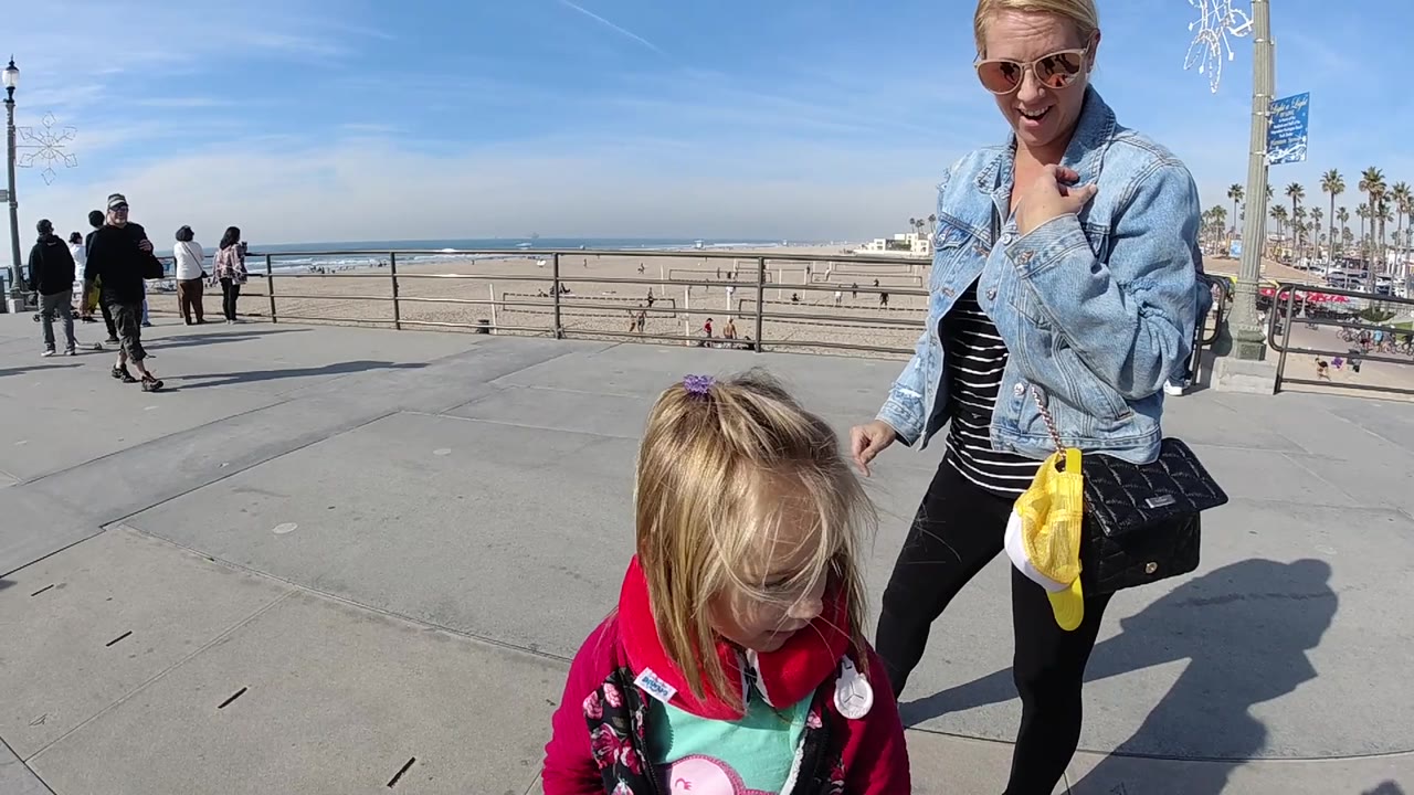 A young child occupies the lower foreground as the woman stands to the right, with a broad Huntington Beach promenade, beach railing, sand, and ocean horizon under clear blue sky.