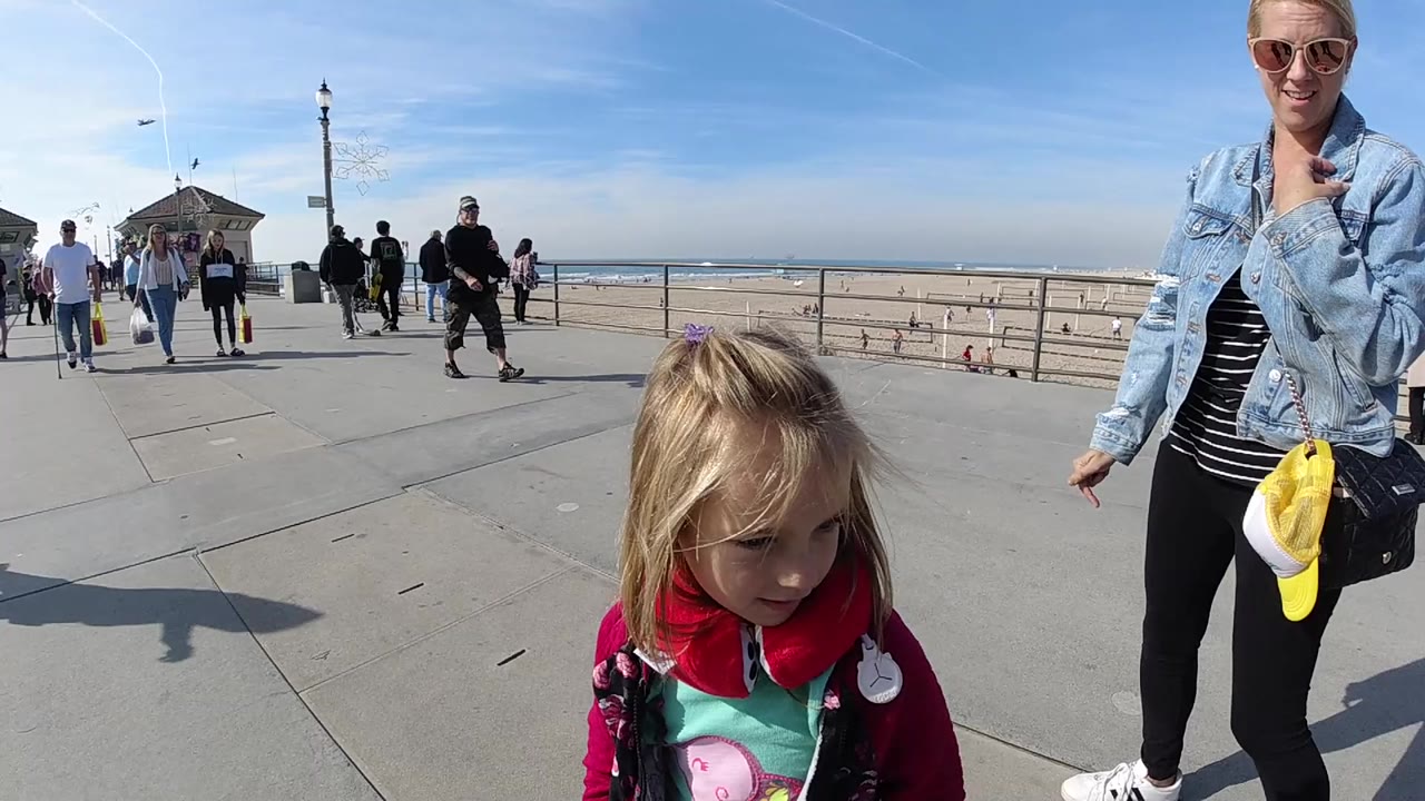 A blond young girl stands in the foreground at Huntington Beach while an adult woman in sunglasses and a denim jacket stands beside her near the beach railing.