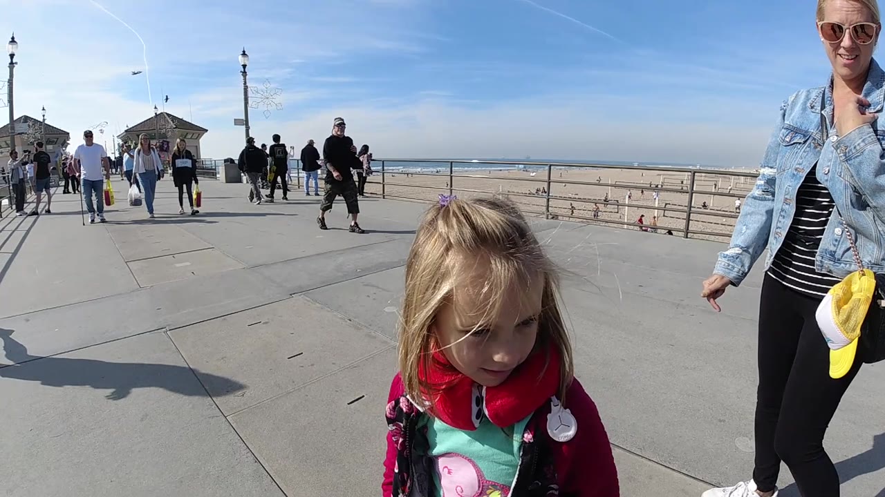 At Huntington Beach, the blond girl looks down in the foreground while an adult woman with a yellow cap at her bag stands partly cropped beside her.