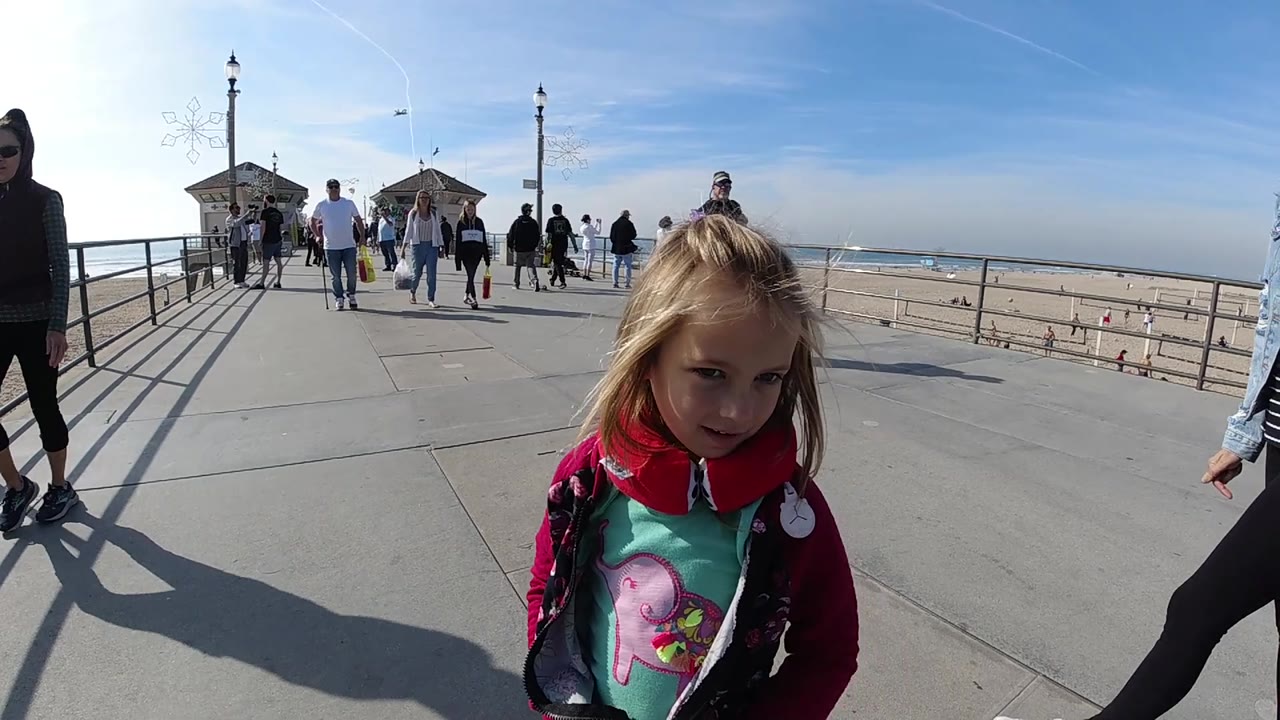 A little girl in a red jacket stands slightly right of center on the Huntington Beach pier, with a wide walkway, rail shadows, distant pedestrians, sand, and ocean behind her.
