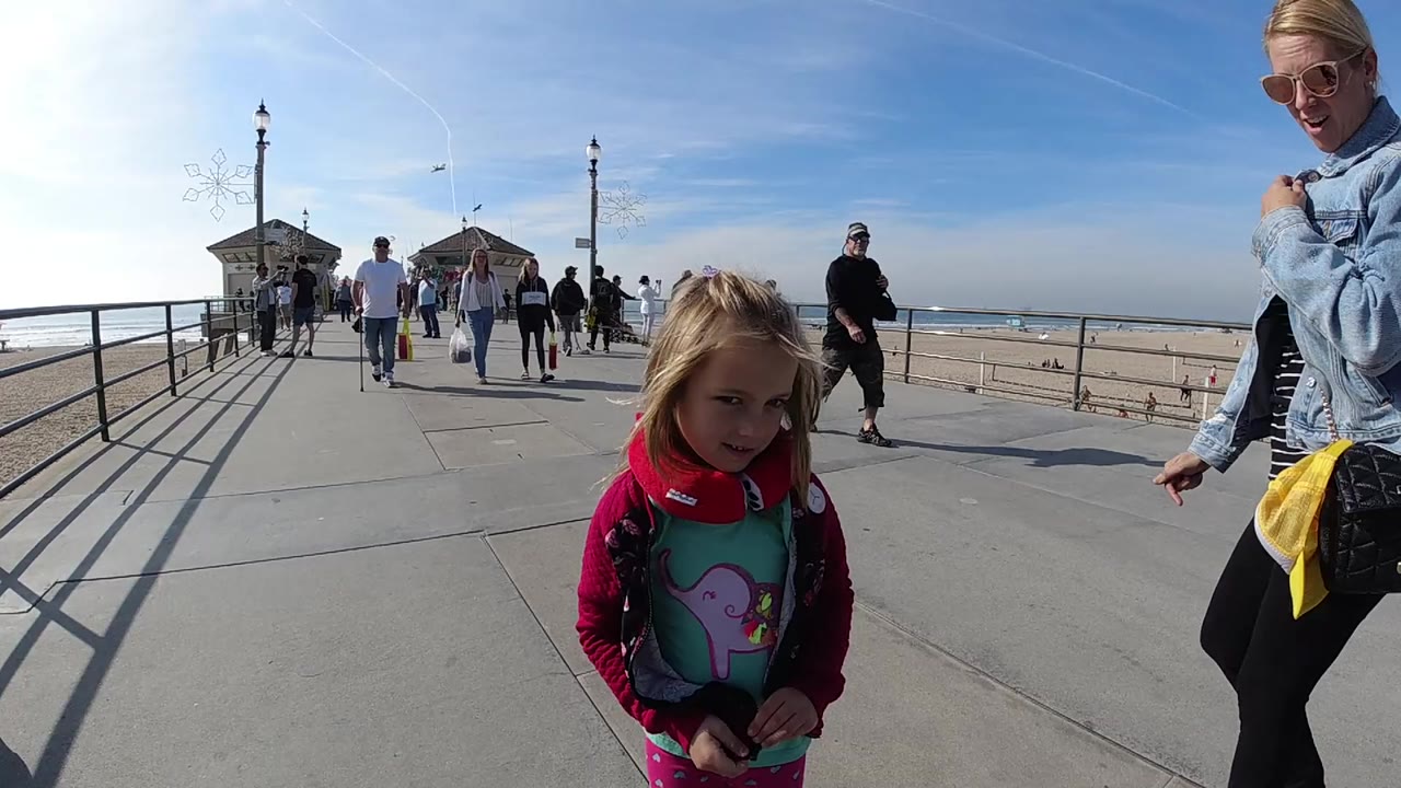 At 0:00 in Huntington Beach, a young blonde girl stands near the foreground while a blonde woman in sunglasses appears at frame right on a sunny pier with pedestrians and ocean behind them.