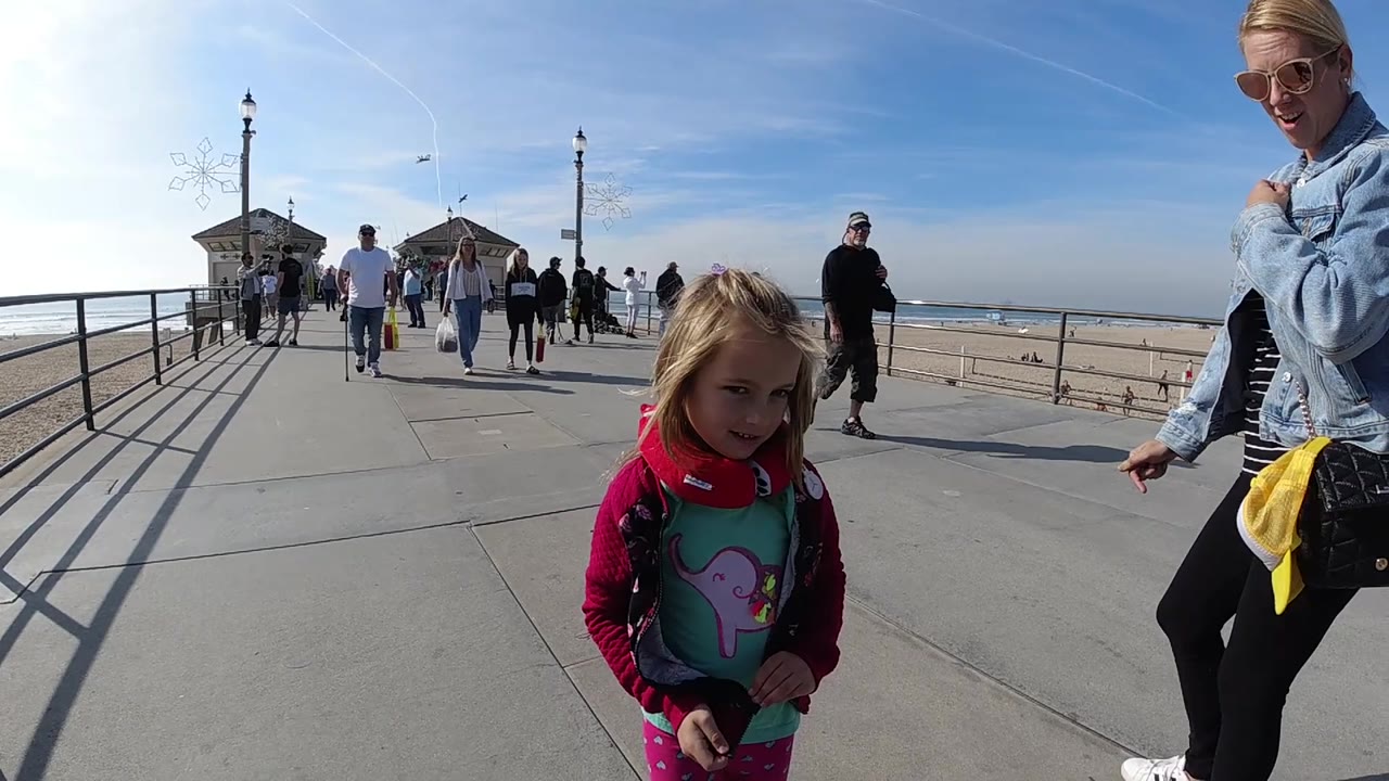 At 0:02 in Huntington Beach, the young girl stands slightly right of center and the woman is cropped along the right edge, with the ocean, beach, railings, and pier buildings visible behind them.