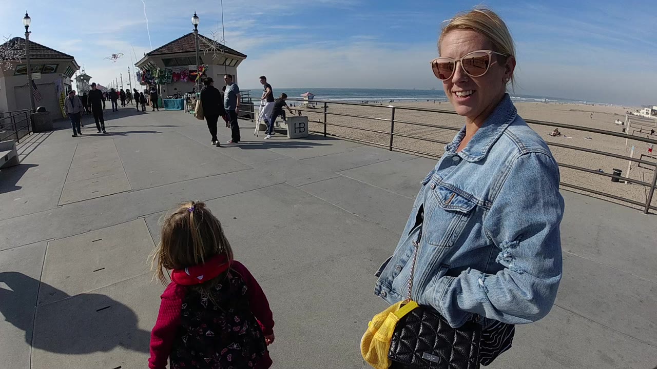 At 0:00 in Huntington Beach, a woman in sunglasses and a denim jacket stands at the right edge while a child in a red hooded top faces the pier walkway and beach.