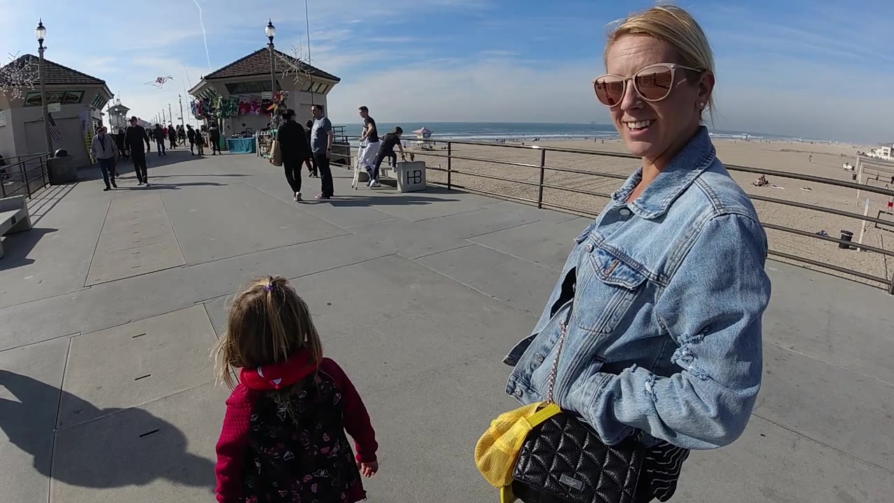 At 0:02 in Huntington Beach, the smiling woman is closest to camera on the right, the child is centered low, and the beach railing leads toward people and pier buildings in the background.
