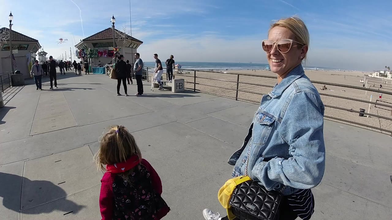 A smiling blond woman in sunglasses stands near a young child on a Huntington Beach pier, with beach sand, ocean horizon, railings, and pedestrians behind them.