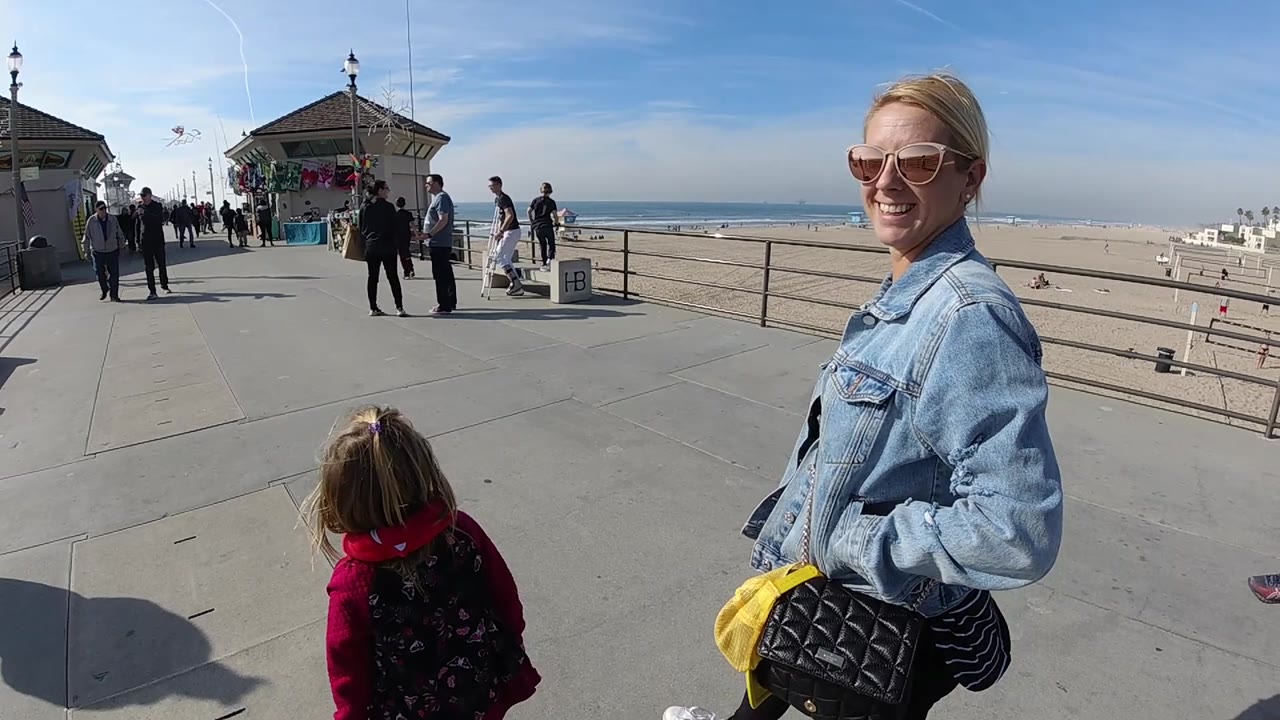 A close foreground view shows the smiling woman and the child from behind on a Huntington Beach pier, with the walkway, railing, beach, and ocean horizon spread across the background.