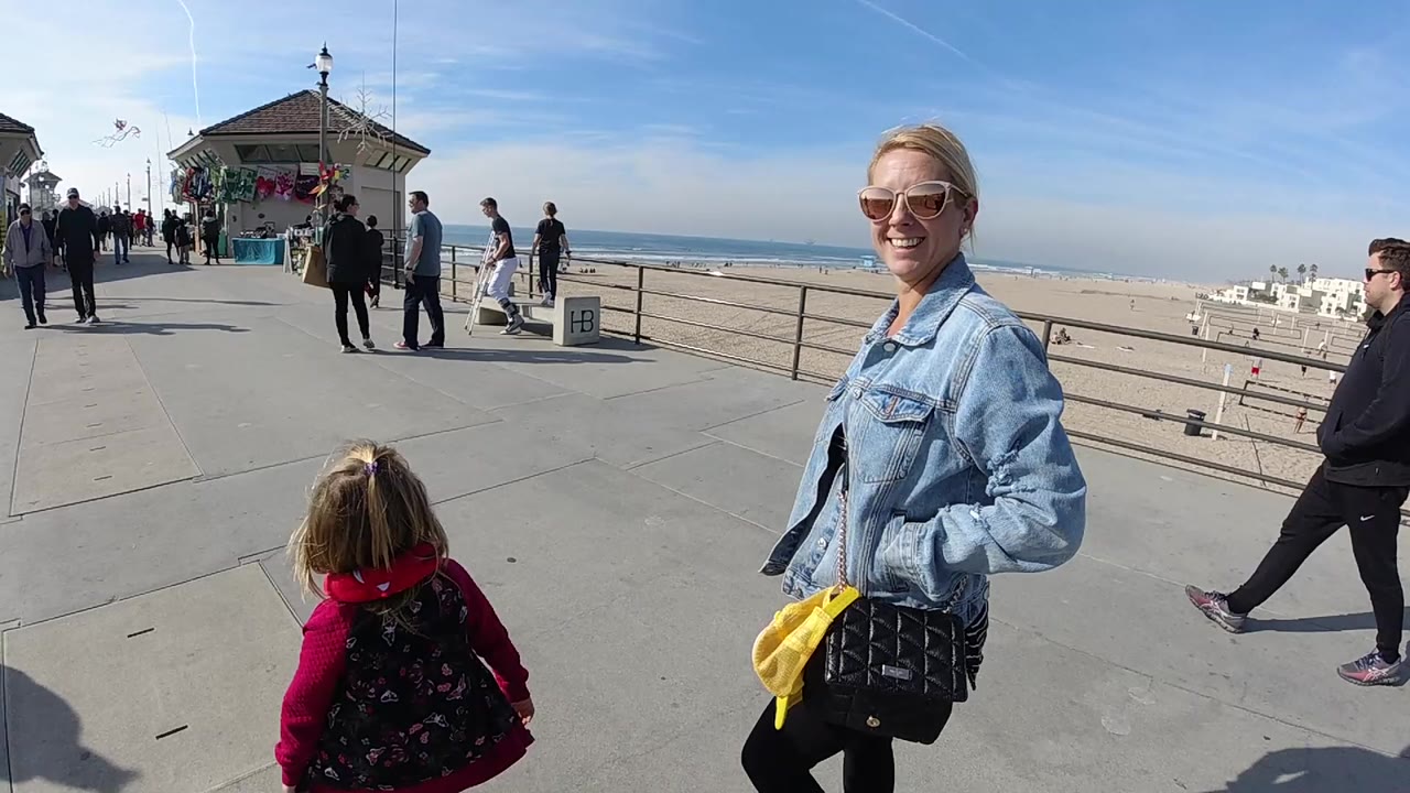 At Huntington Beach, California, a smiling woman in sunglasses stands near the camera while a child in red faces the pier walkway, with pedestrians, sand, ocean, and blue sky behind them.