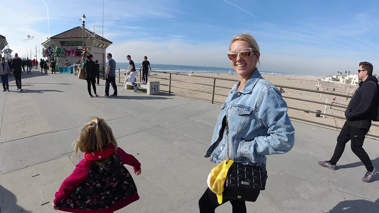 The child shifts farther across the foreground at Huntington Beach as the woman smiles near the right edge, with walkers, railings, the sandy beach, and the ocean horizon filling the background.