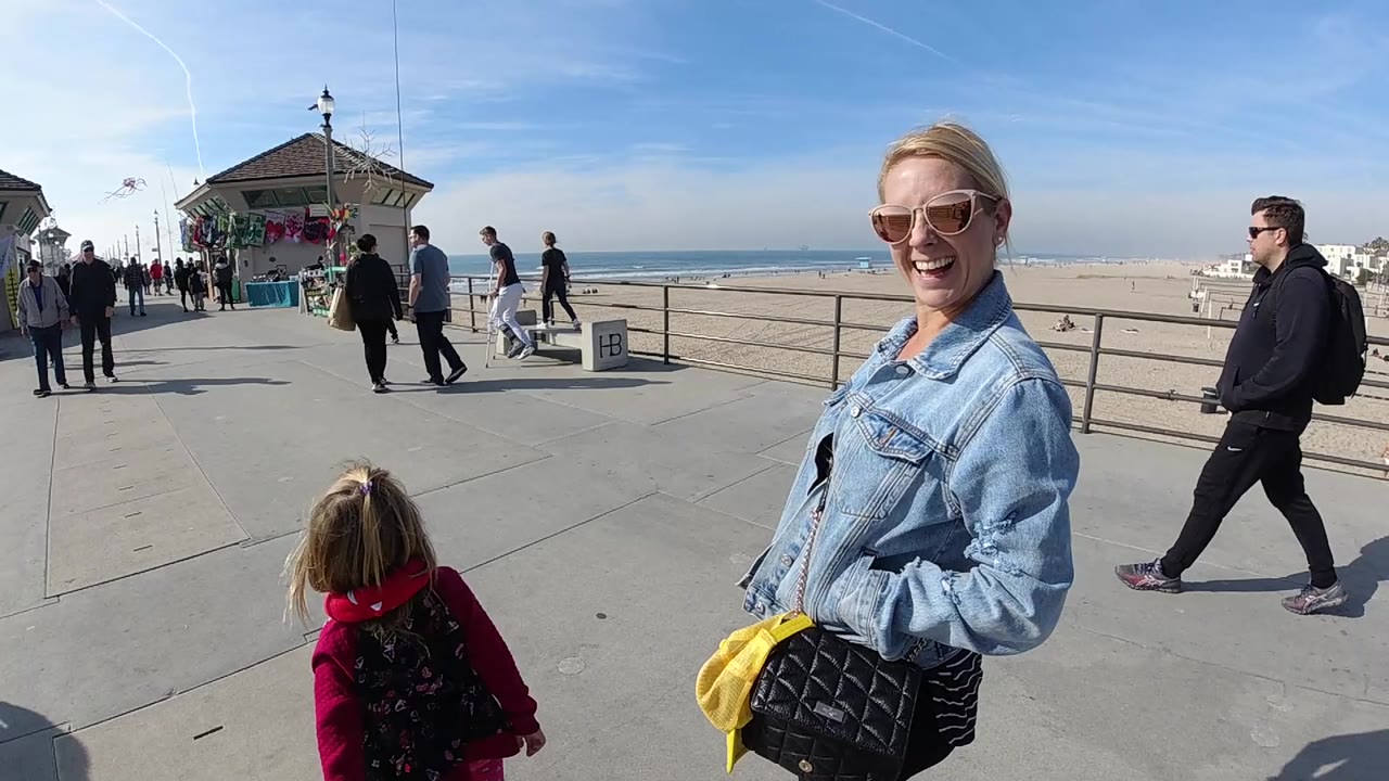 Child in a red jacket walks forward as a woman smiles near the camera by the beach.