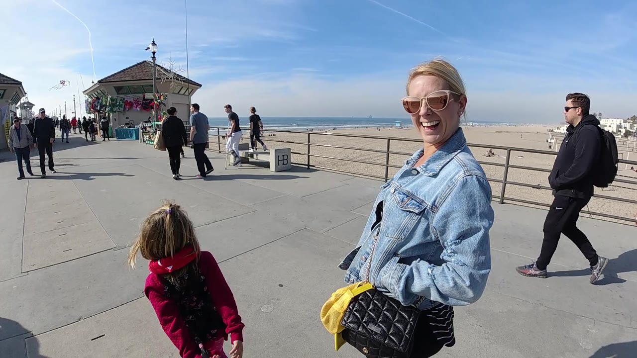 At 0:02 in Huntington Beach, California, the woman in a denim jacket smiles near the right edge as the girl in red continues along the walkway with the beach building and ocean behind.