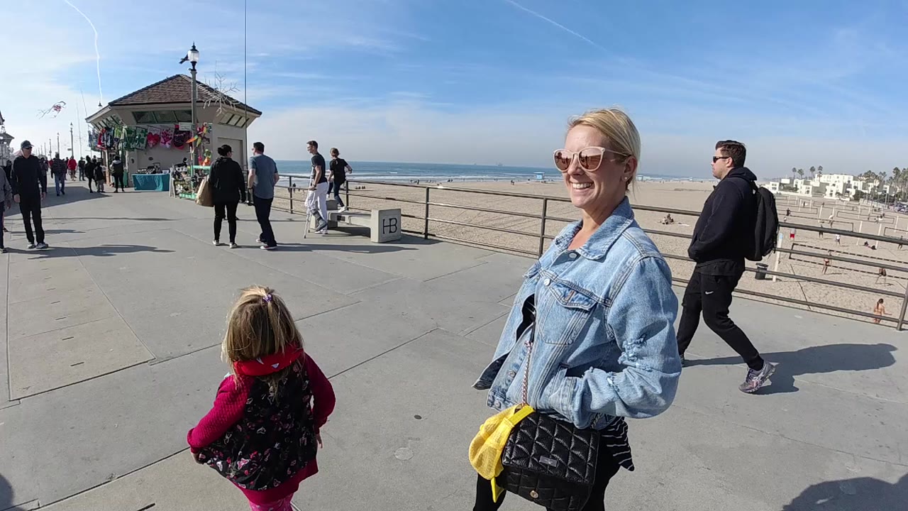 In Huntington Beach, a smiling woman in sunglasses and a light denim jacket stands near a child in pink on a paved boardwalk with ocean, railings, pedestrians, and a small beach structure behind them.