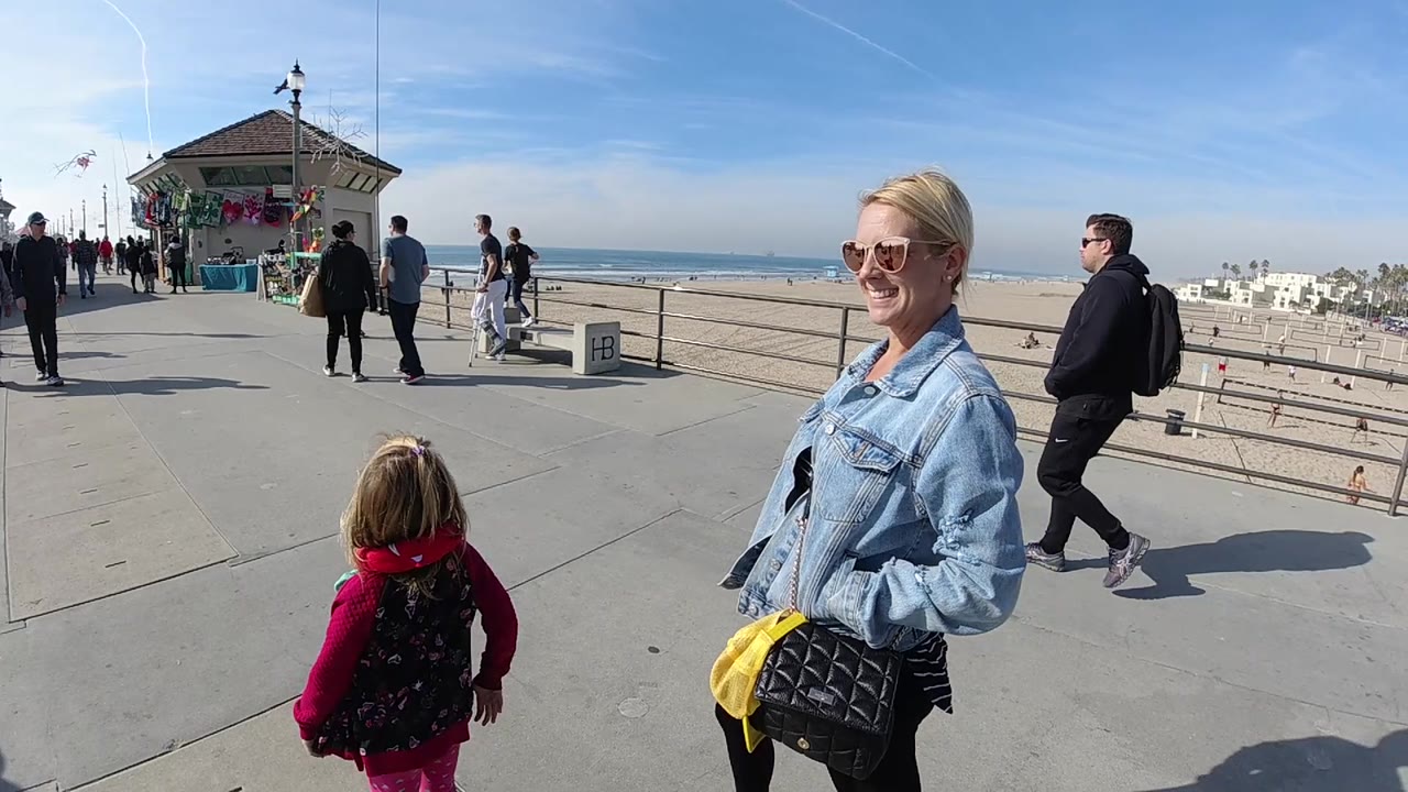 In Huntington Beach, the child in pink is centered lower in frame while the woman in denim and sunglasses stands to the right, with walkers, railings, ocean, sand, and distant beach buildings in sunlit background.