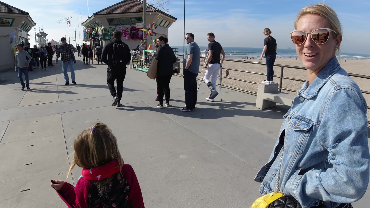 At 0:00 in Huntington Beach, California, a woman in sunglasses and a denim jacket stands near camera while a child in a red jacket walks ahead on a crowded beachfront walkway.